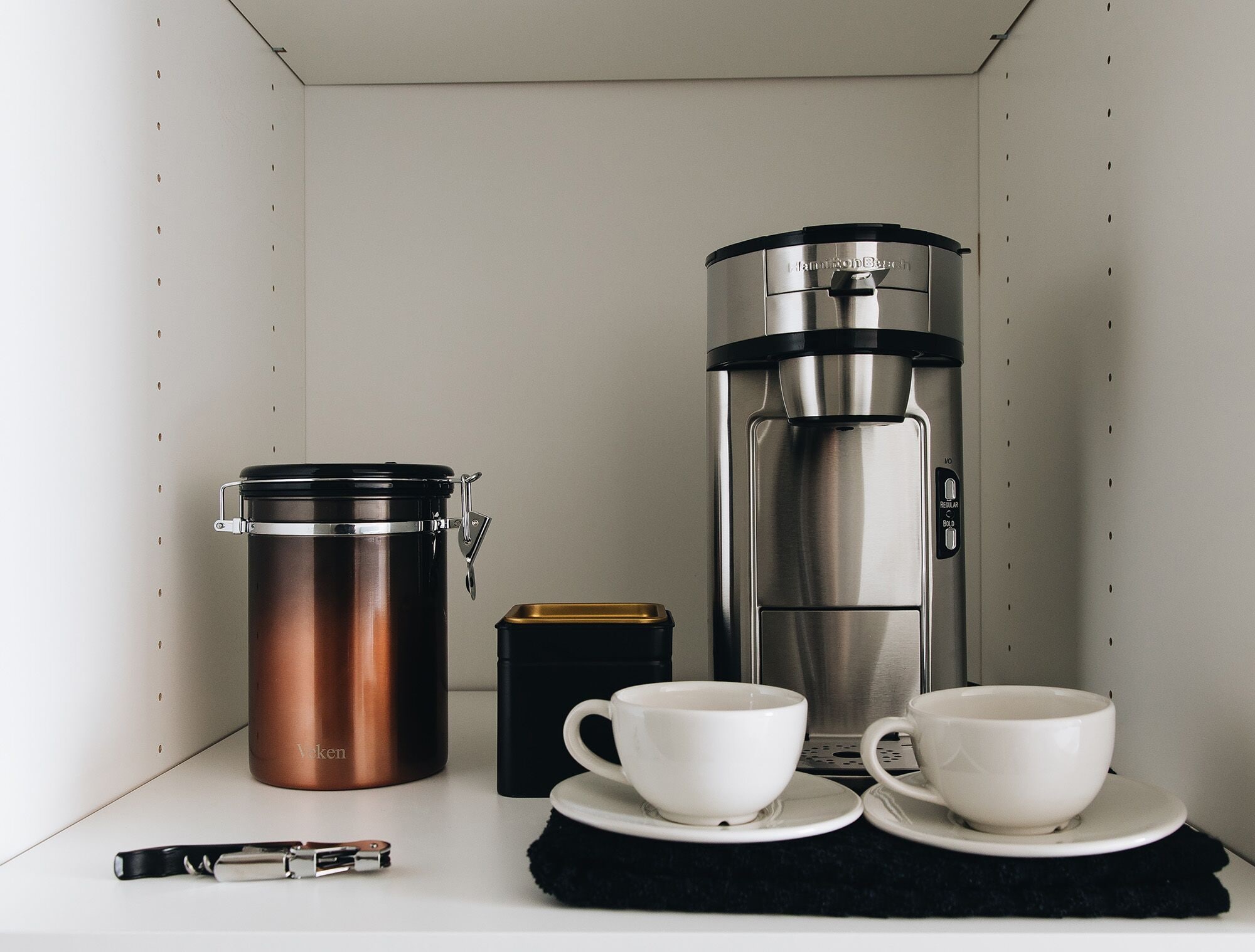 A kitchenette with a coffee maker, a thermos, a box, and two cups on a countertop.
