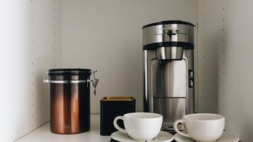 A kitchenette with a coffee maker, a thermos, a box, and two cups on a countertop.