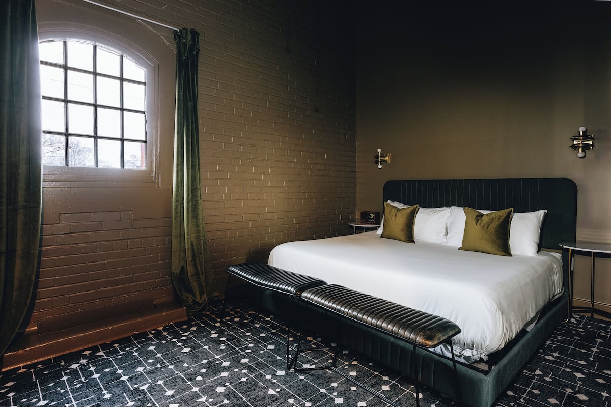 A hotel room with a large bed, a bench, a small table, and a window with curtains.