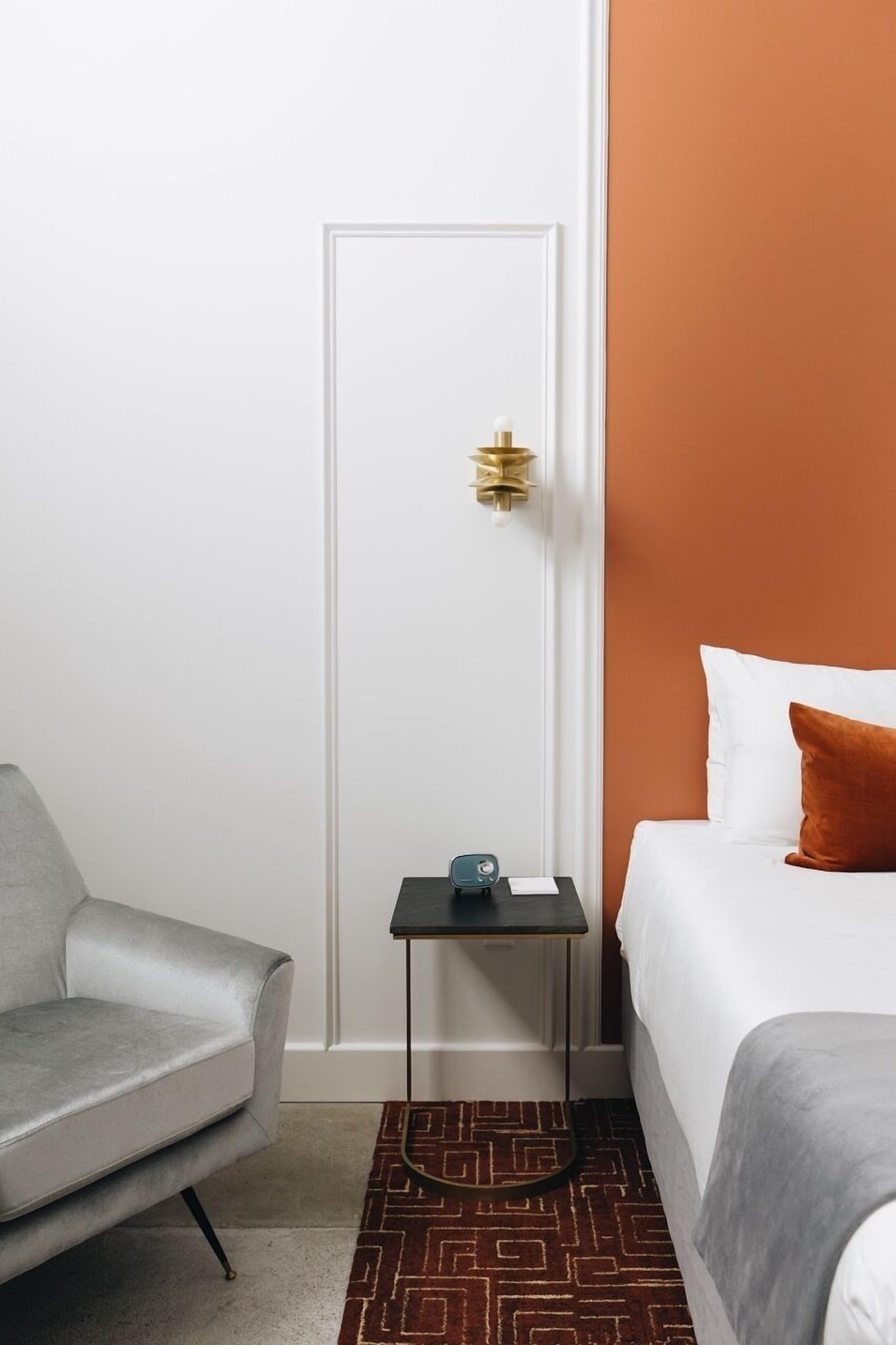 A hotel room with a white door, a small table with a lamp, a grey sofa, and a bed with white and orange bedding.