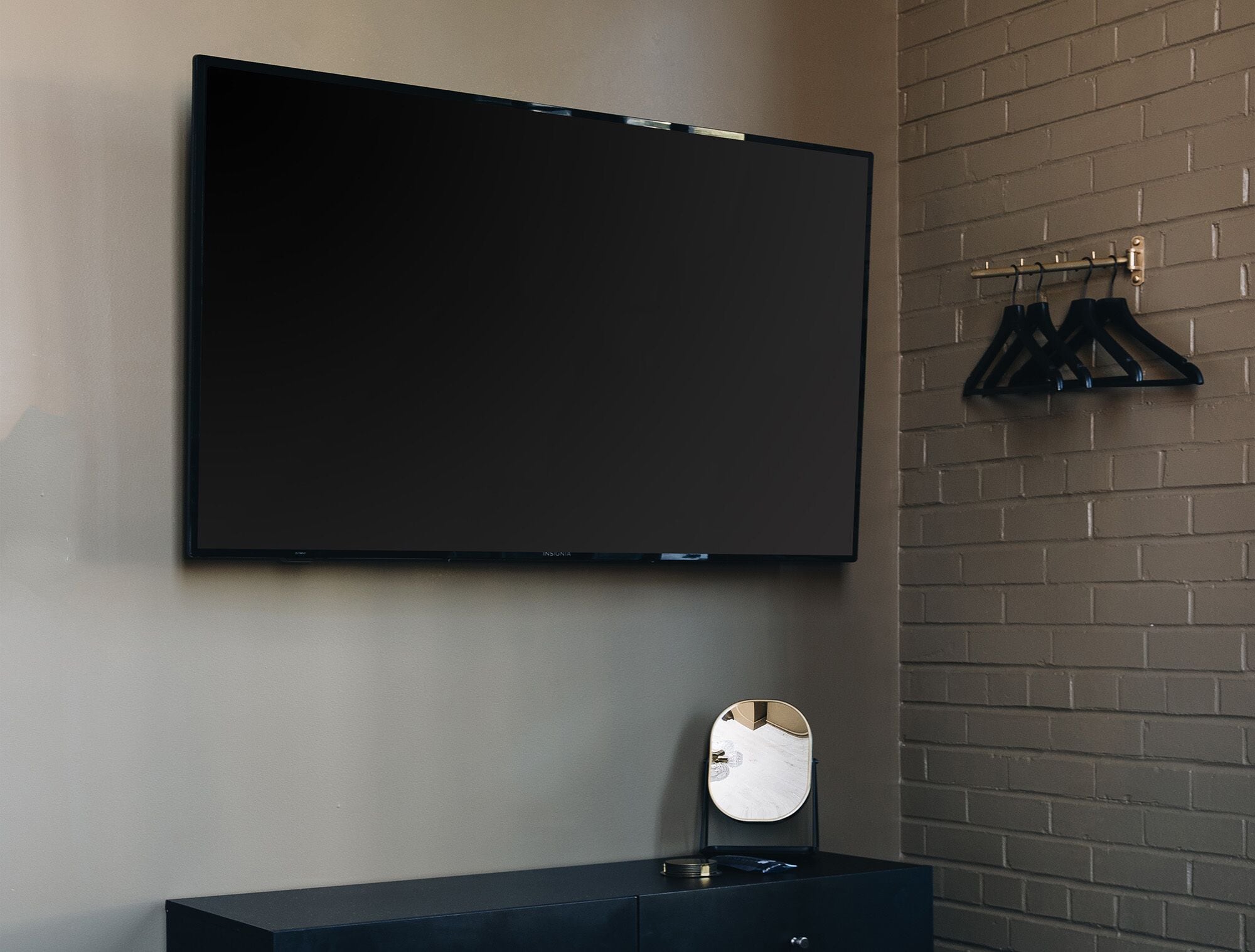 Una habitación moderna con un televisor de pantalla plana grande montado en la pared, un espejo pequeño sobre una cómoda oscura, y una pared con diseño de ladrillo.