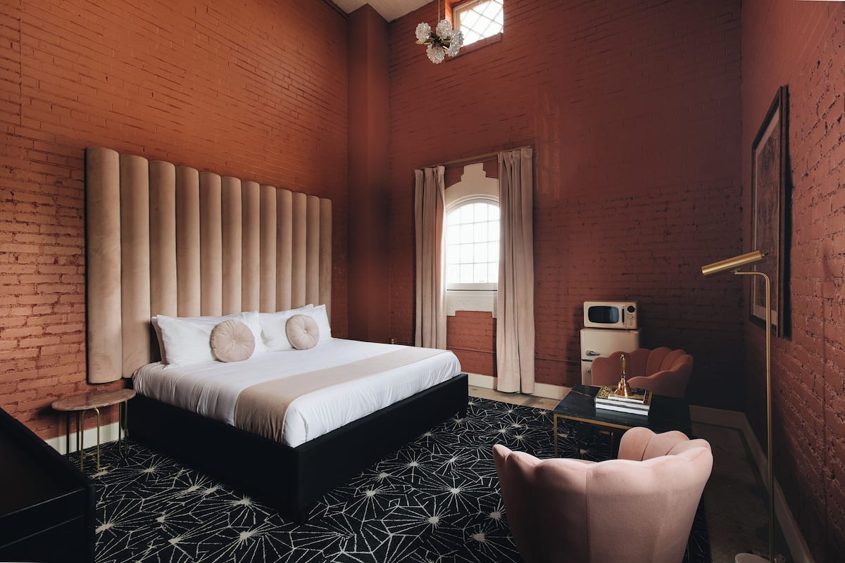 A modern bedroom with a brick wall, a large bed with a headboard, a small round table, a chair, and a TV.