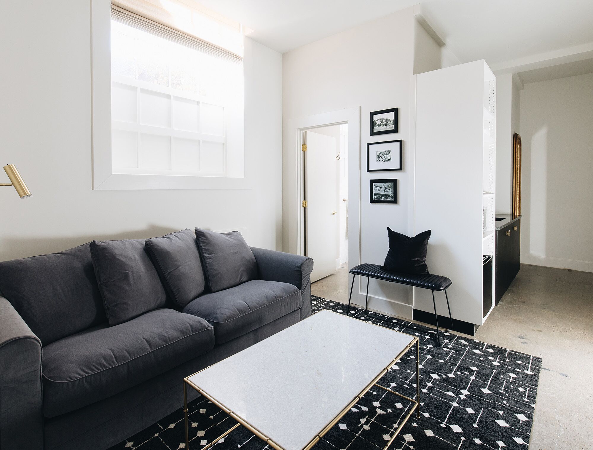 A modern living room with a grey sofa, a small table, and a patterned rug. There is a door leading to another room and a wall with framed pictures.