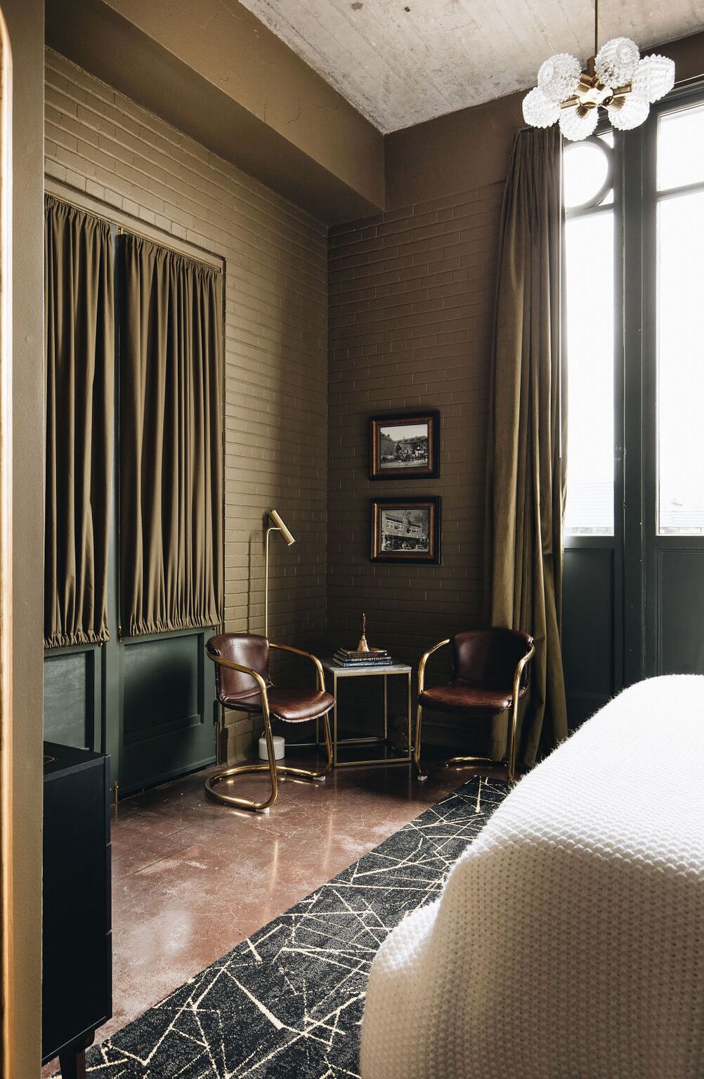 Una habitación de hotel con pared de ladrillo, dos cuadros enmarcados, una pequeña mesa con lámpara, dos sillones de cuero y una cama con cubrecama blanco.