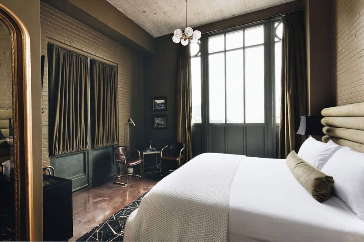 A hotel room with a large bed, a desk, two chairs, a window with curtains, and a chandelier.