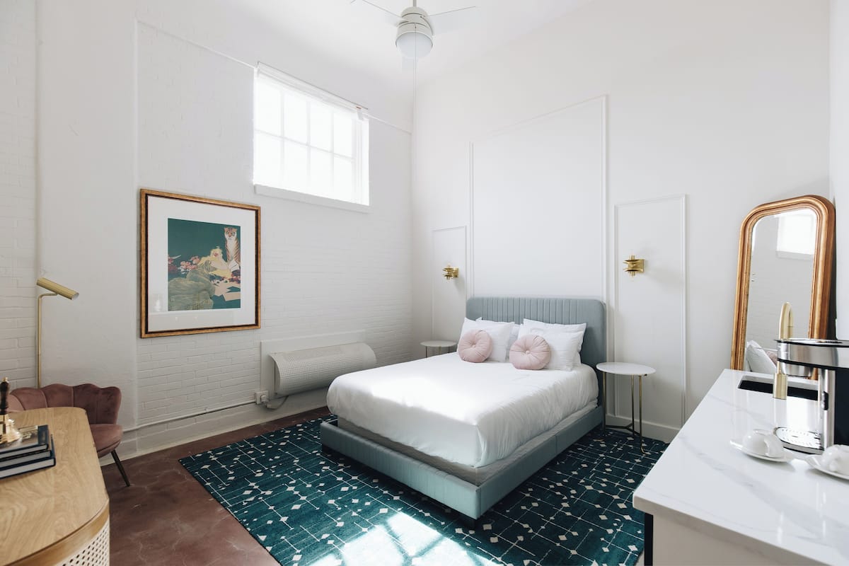 A bedroom with a bed, a bedside table, a chair, a desk, a mirror, a window, and a picture on the wall.