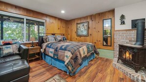 Een gezellige slaapkamer met een groot bed, een houten vloer, een open haard en een raam met uitzicht op groen.