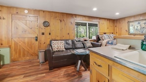 Een gezellig interieur van een chalet met een houten gevel, een grote schuifdeur, een leren bank, een kleine tafel en een keuken met een spoelbak en kastjes.