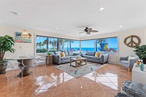 A spacious living room with a large window overlooking a beach, a ceiling fan, and a view of palm trees.
