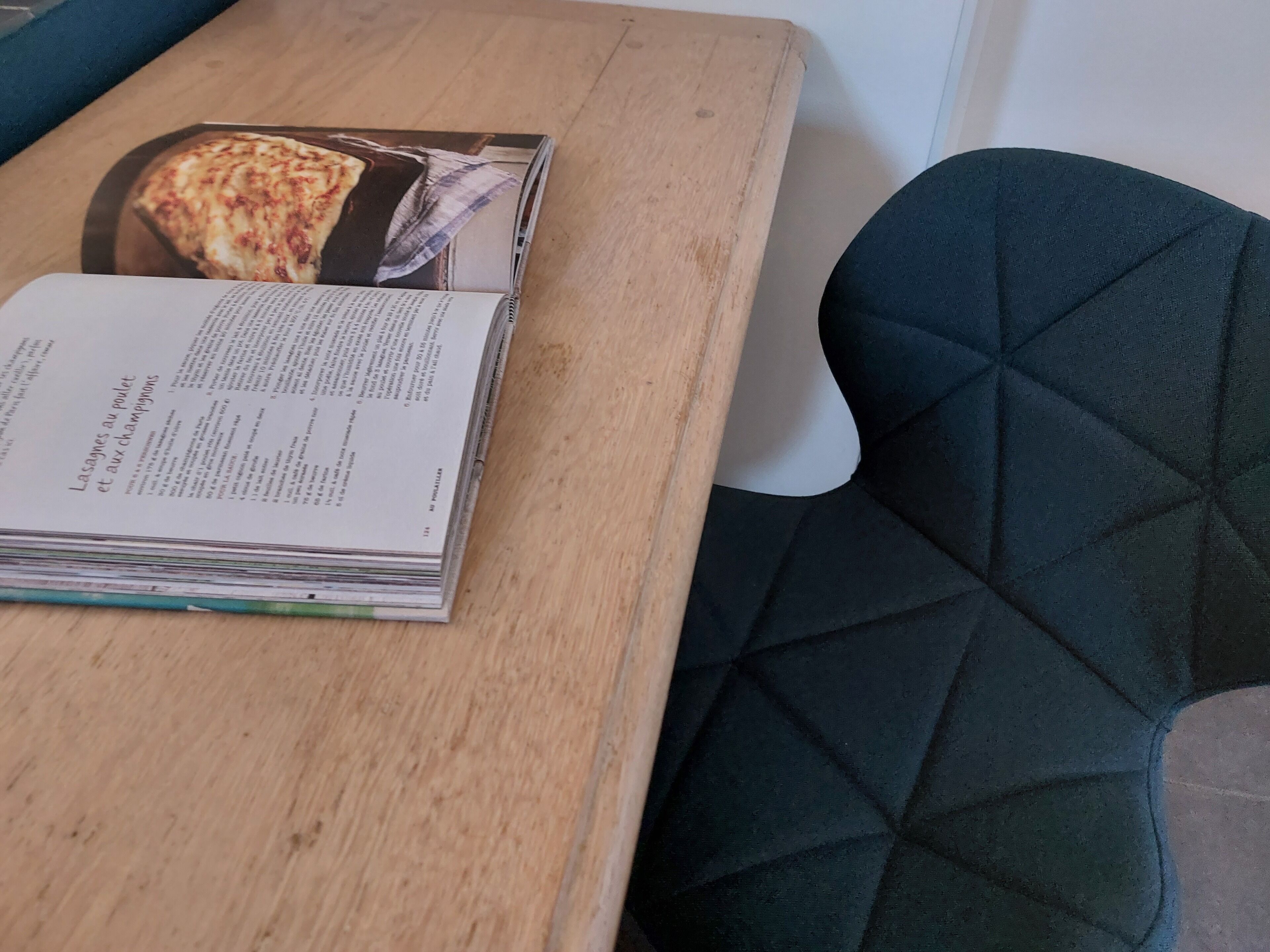 Een boek met de titel 'Lasagnes au poulet et aux champignons' op een houten tafel, naast een donkerblauwe fauteuil.