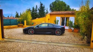 Une voiture de sport noire est garée dans une allée, avec une maison jaune et un mur de pierres en arrière-plan.
