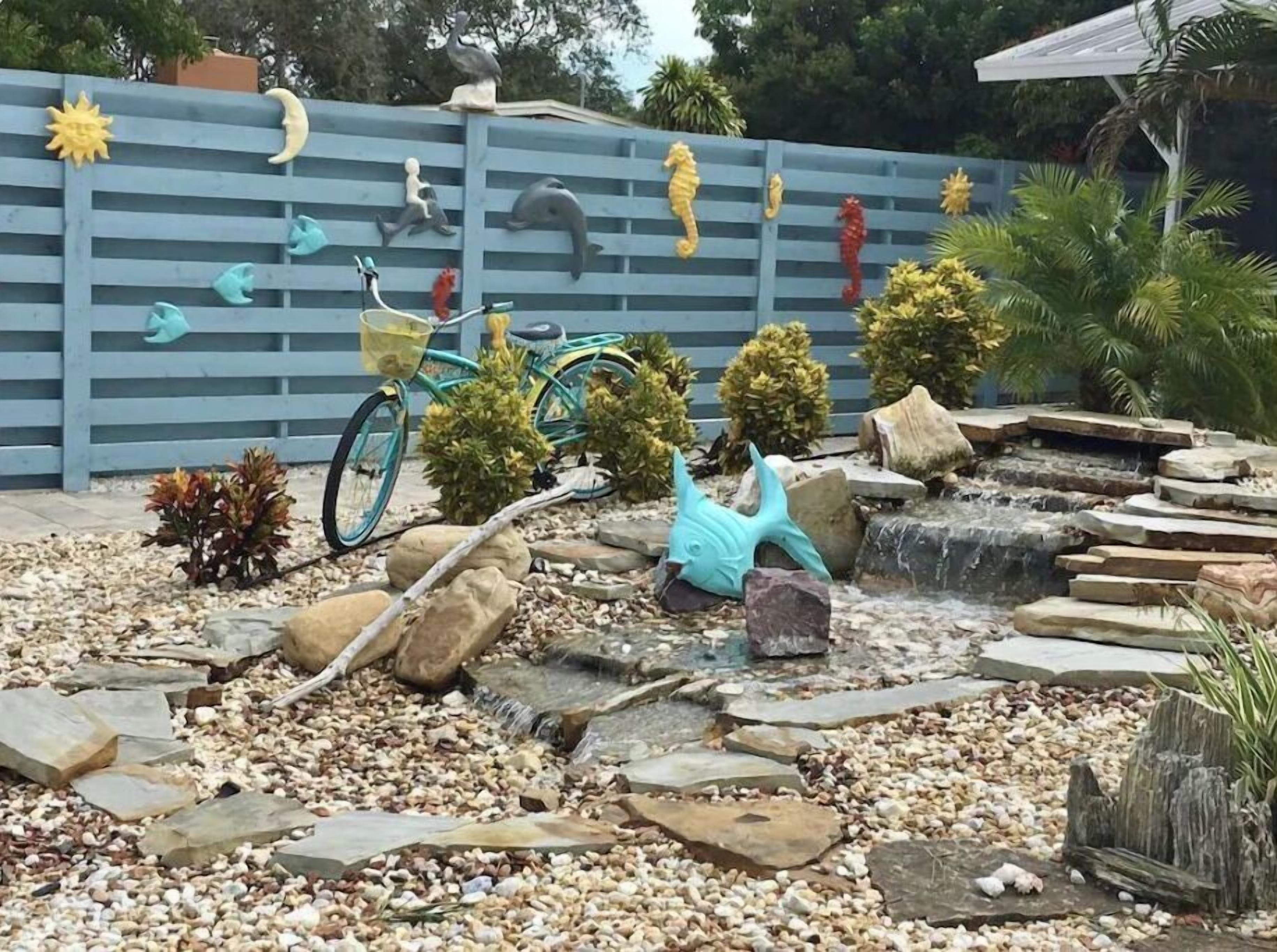 Un jardín con valla azul, seahorses decorativos, una bicicleta y una pequeña cascada.