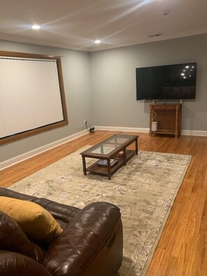 A living room with a brown leather couch, a glass coffee table, a patterned rug, a flat-screen TV, and a wooden cabinet.