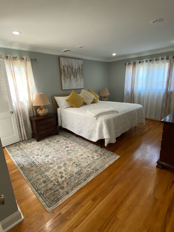 A bedroom with a double bed, two bedside tables with lamps, a large window with curtains, and a patterned rug.