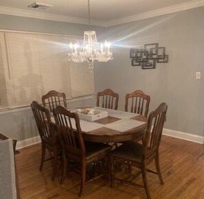 A dining room with a round wooden table, six wooden chairs, a chandelier, and a window with blinds.