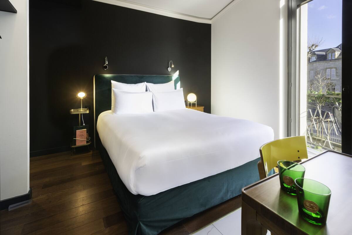 Ein Hotelzimmer mit einem großen Bett, Nachttischen mit Lampen, Blick durch ein Fenster nach draußen und einem kleinen Tisch mit Stuhl.