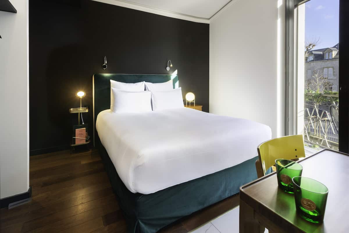 Ein Hotelzimmer mit Bett, Nachttischlampen, einem Tisch mit Gläsern und Blick auf Gebäude.