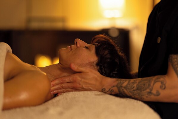 A person lying on a bed with a blanket, another person with tattoos on their arm is touching their shoulder.