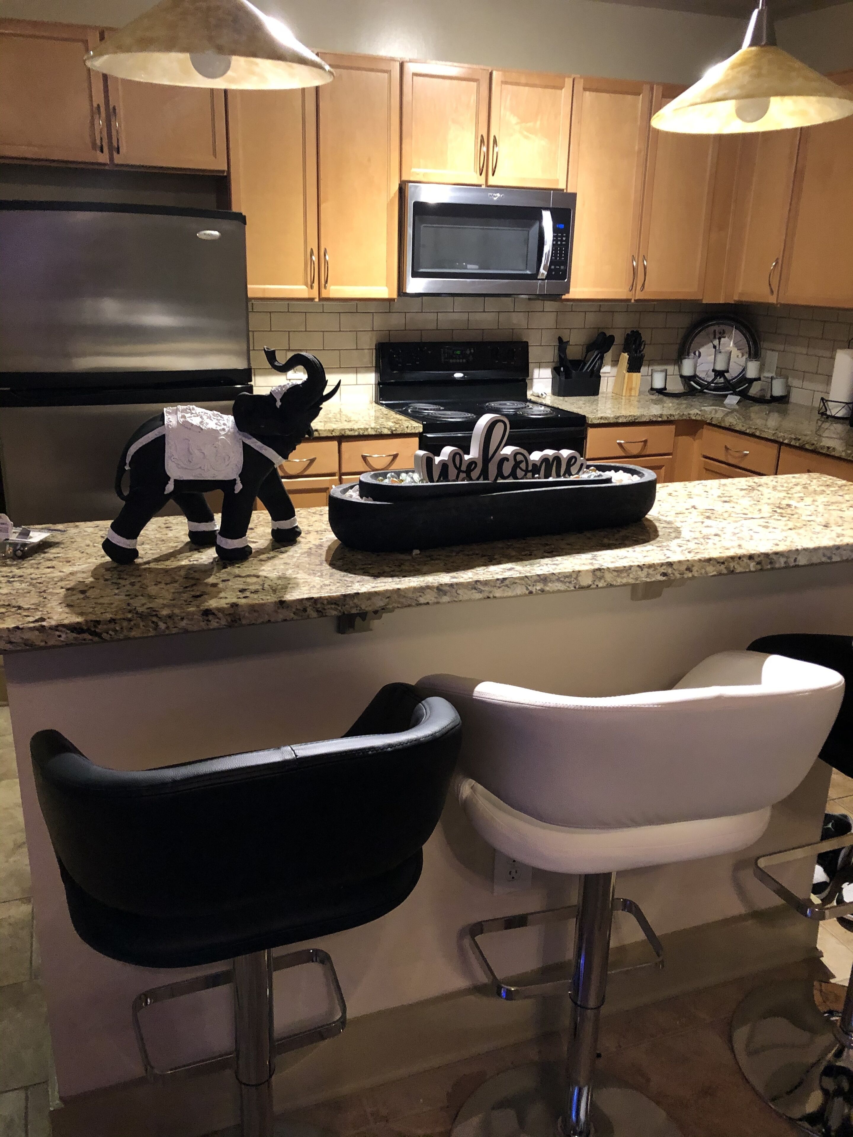 Una cocina con armarios de madera, un refrigerador de acero inoxidable, un microondas y una estufa. Hay dos taburetes de bar y una figurita decorativa de elefante.