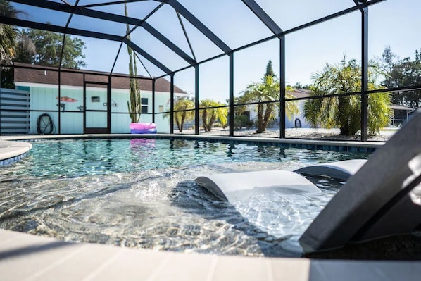 A swimming pool with a hot tub area, surrounded by a glass enclosure, and palm trees in the background.