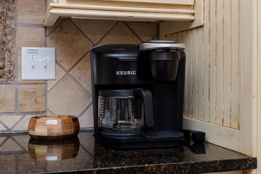 Keurig-kahvinkeitin keittiön työtasolla, sen vieressä puinen kulho, jossa on merkintä ”K-kahvikapselit”, ja roskasäiliö.