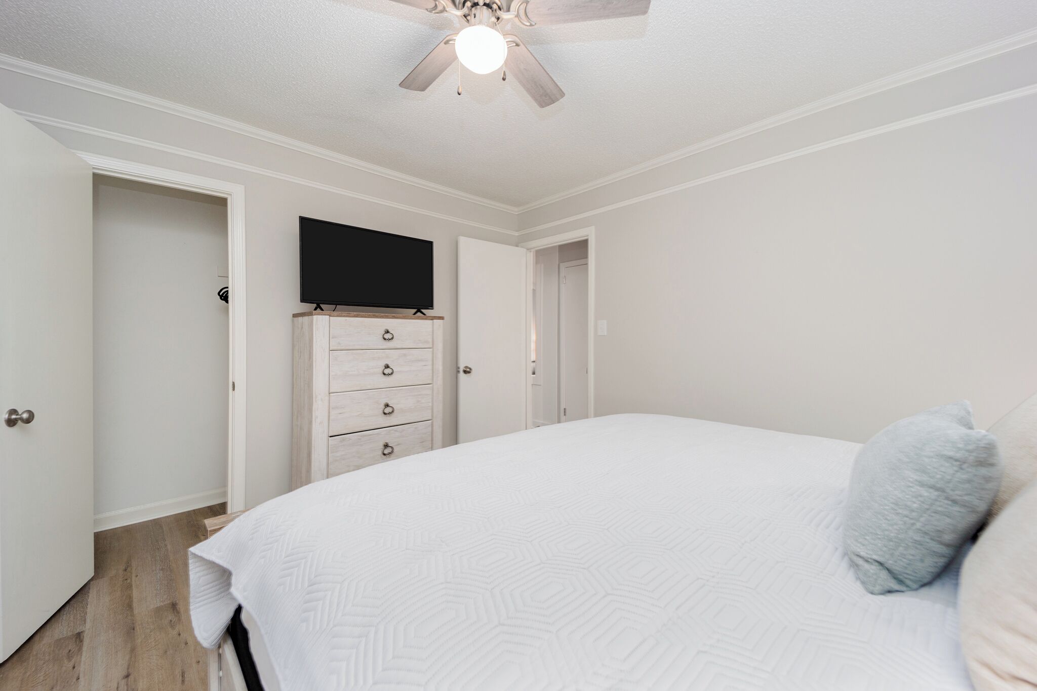 A bedroom with a bed, a ceiling fan, a TV mounted on a dresser, and a door leading to another room.
