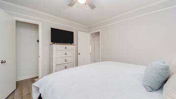 A bedroom with a bed, a ceiling fan, a TV mounted on a dresser, and a door leading to another room.