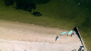 Vue aérienne d’une plage aux eaux cristallines, bordée d’un rivage sablonneux, où l’on aperçoit plusieurs kayaks et planches à pagaie. Des chaises longues et des parasols sont disposés le long de la plage.