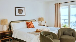 Une chambre à coucher avec un lit, deux tables de chevet, une chaise, une fenêtre donnant sur un paysage agréable et un tableau encadré au mur.