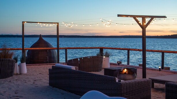 Une terrasse au bord de la plage, avec des meubles en osier, un pouf blanc et un foyer.
