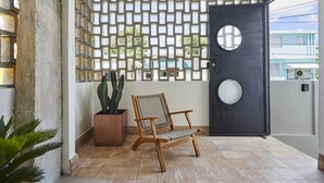 Una silla de madera frente a una puerta negra con un ventanal circular, un cactus en una maceta y un muro con diseño.