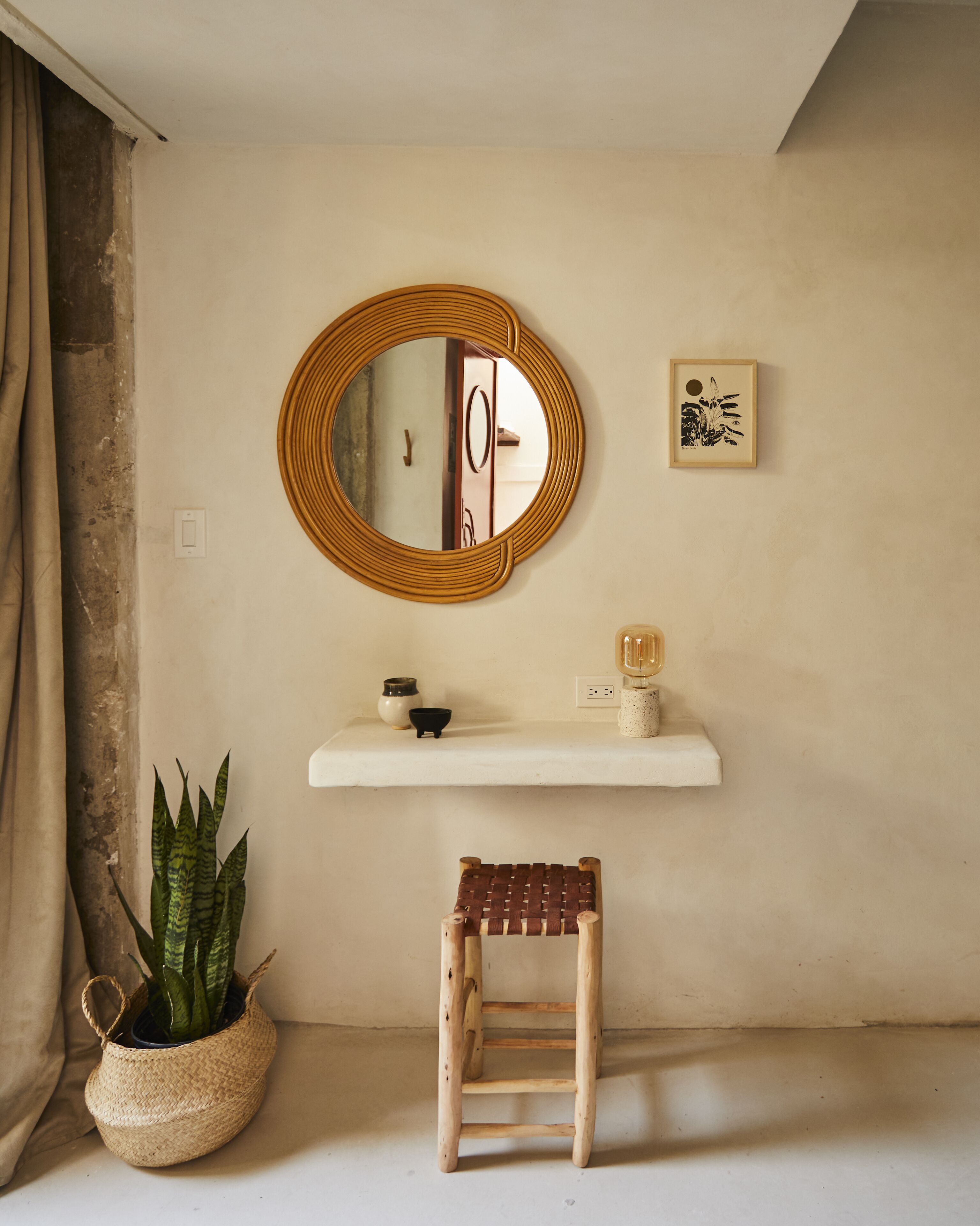 A room with a round mirror, a small shelf with decorative items, a woven stool, and a potted plant.