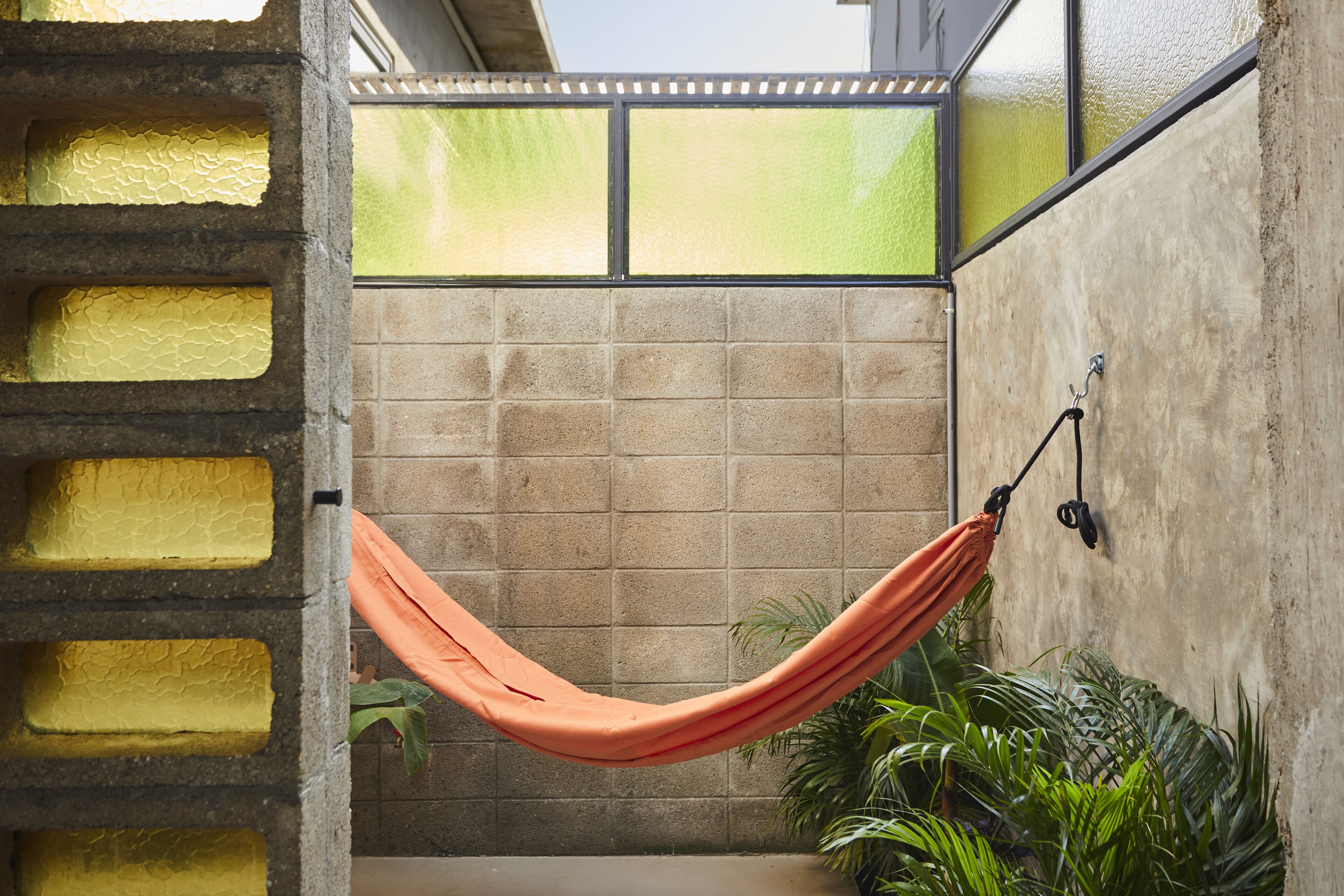 A hammock suspended between a concrete wall and a glass partition, with a view of a tiled floor and green plants.