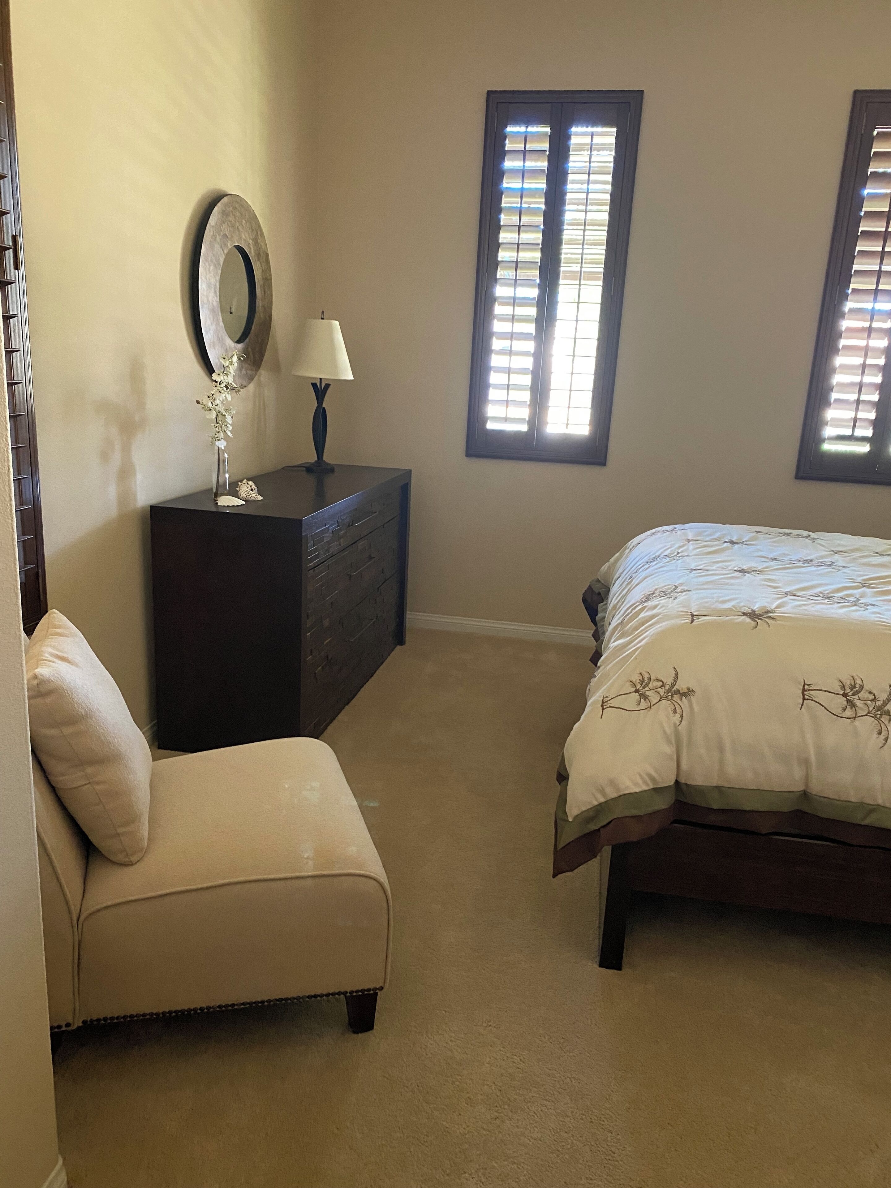 Un dormitorio con cama, una silla, cómoda, espejo y dos ventanas.