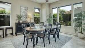 A dining area with a round table, six chairs, a ceiling fan, and large windows offering views of greenery.