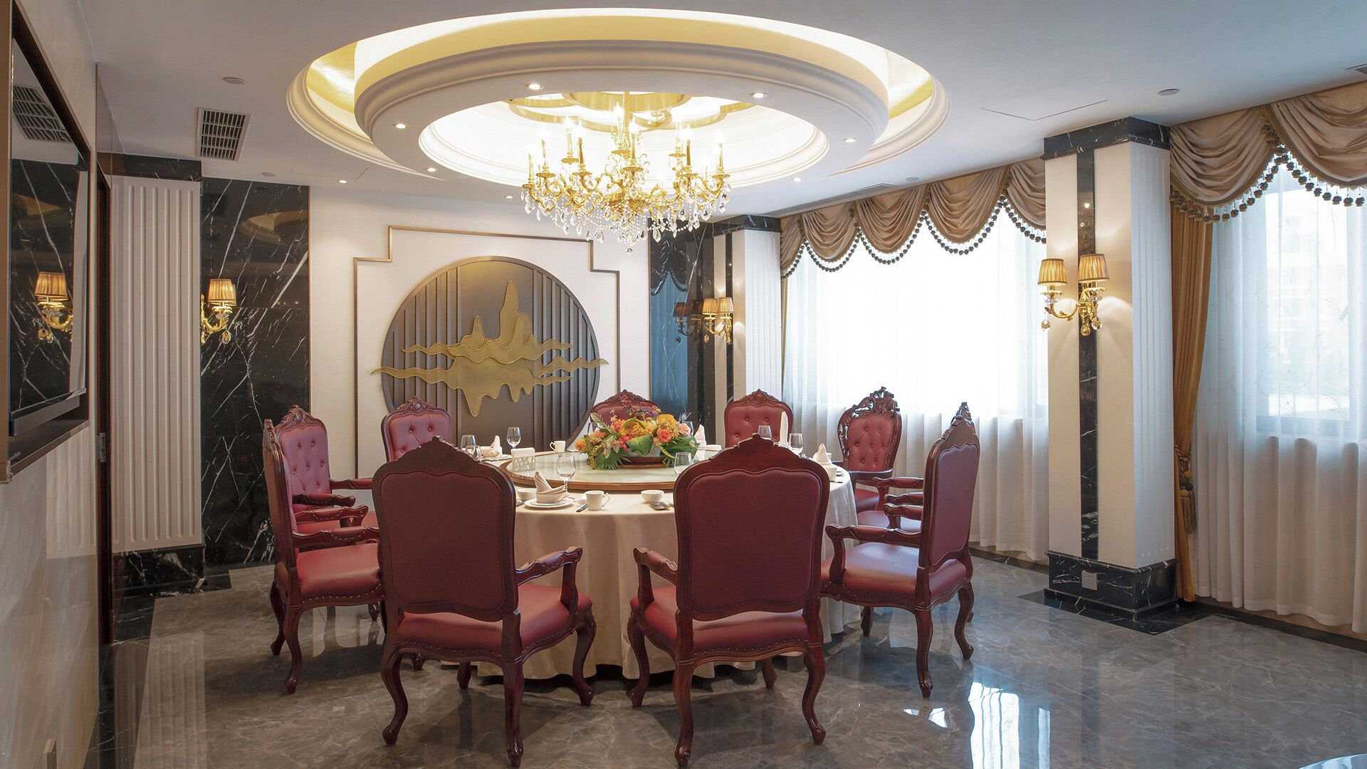 A formal dining room with a round table, six upholstered chairs, a chandelier, and a decorative wall piece.