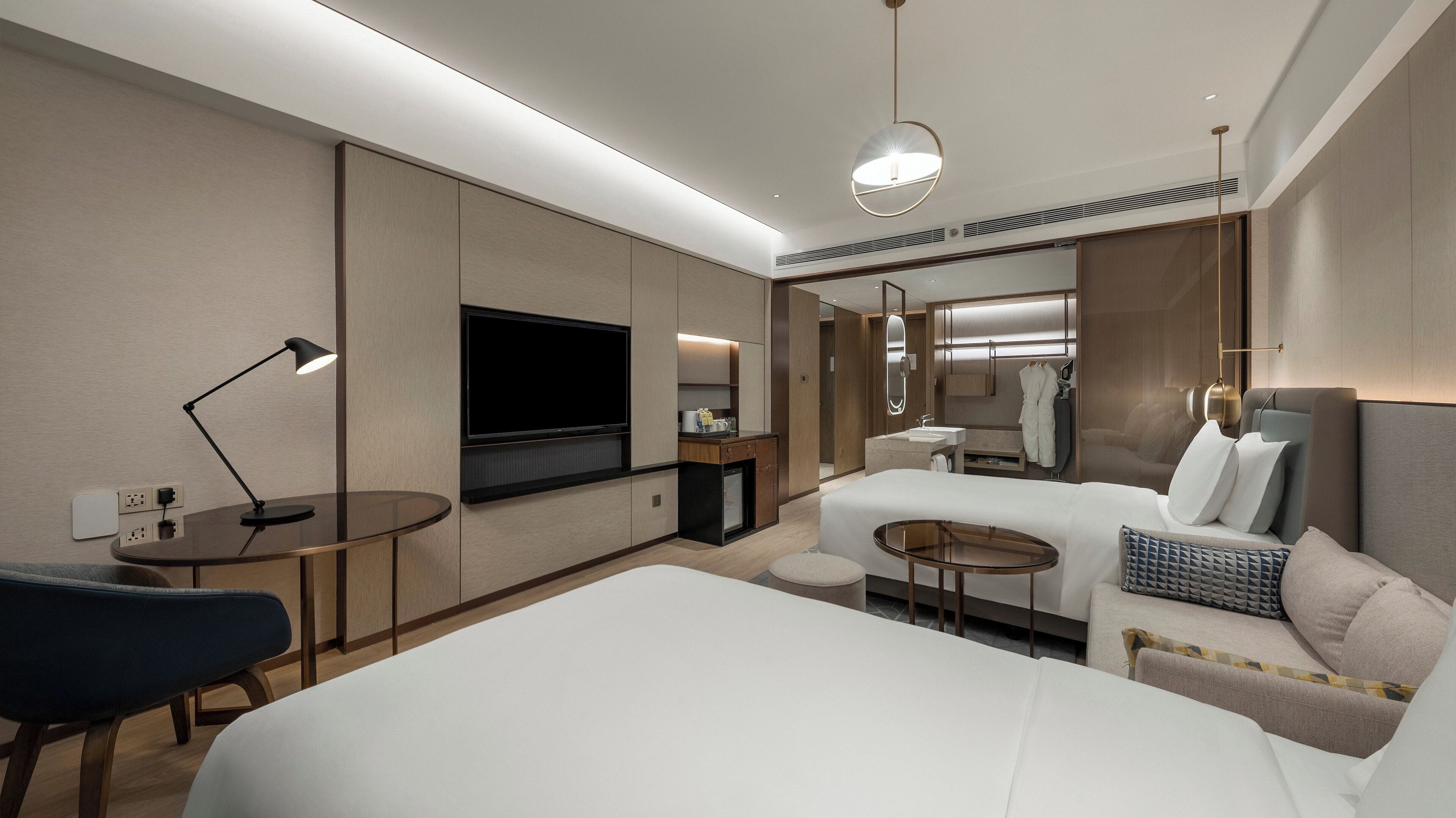 Ein modernes Hotelzimmer mit einem großen Bett, einem Schreibtisch mit Lampe, einem Fernseher, einem kleinen Tisch und einem Badezimmer mit Spiegel und Waschbecken.