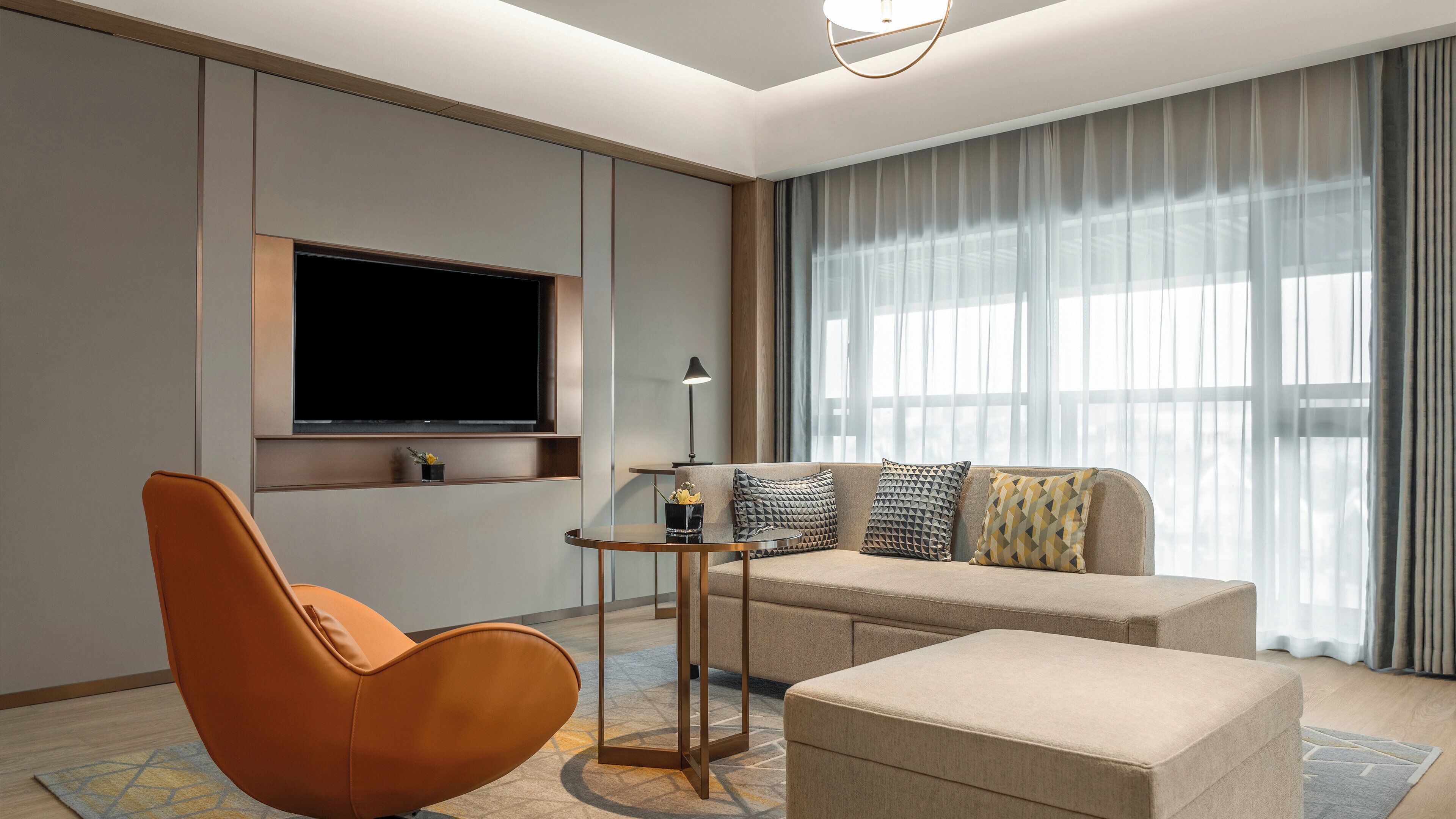Ein modernes Wohnzimmer mit einer Couch, einem Hocker und einem einzelnen Sessel. Ein Flachbildfernseher ist an der Wand befestigt, und ein kleiner Tisch mit einer Lampe steht daneben.