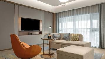 Ein modernes Wohnzimmer mit einer Couch, einem Hocker und einem einzelnen Sessel. Ein Flachbildfernseher ist an der Wand befestigt, und ein kleiner Tisch mit einer Lampe steht daneben.
