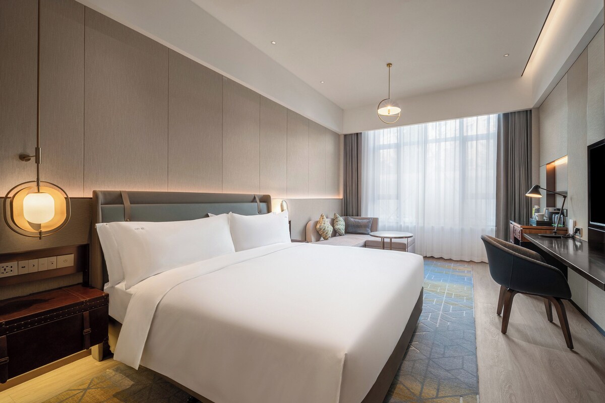 A hotel room with a large bed, a desk, a chair, a TV, and a window with curtains.