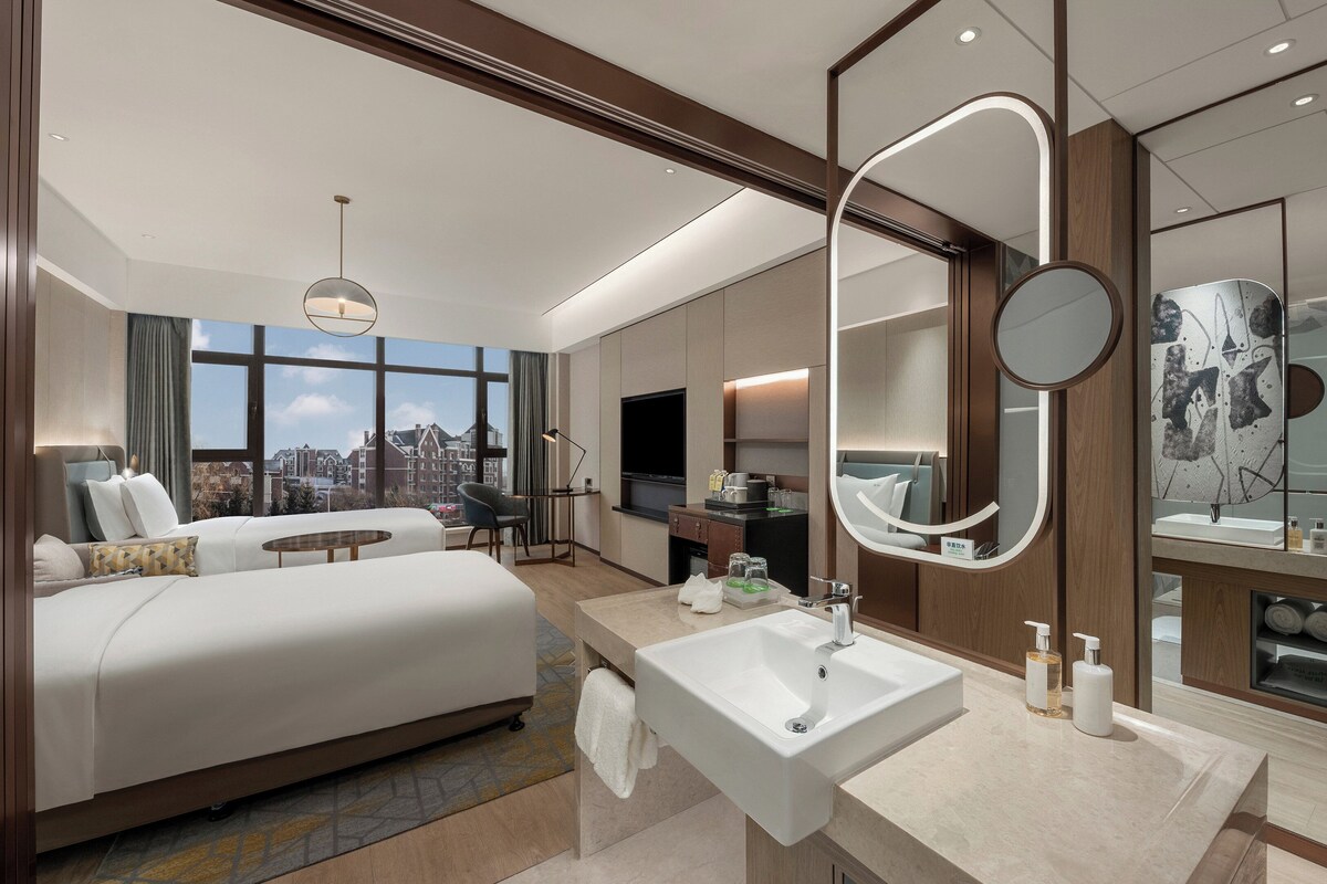 A modern hotel room with a large bed, a sitting area, a TV, a bathroom with a mirror, sink, and shower.