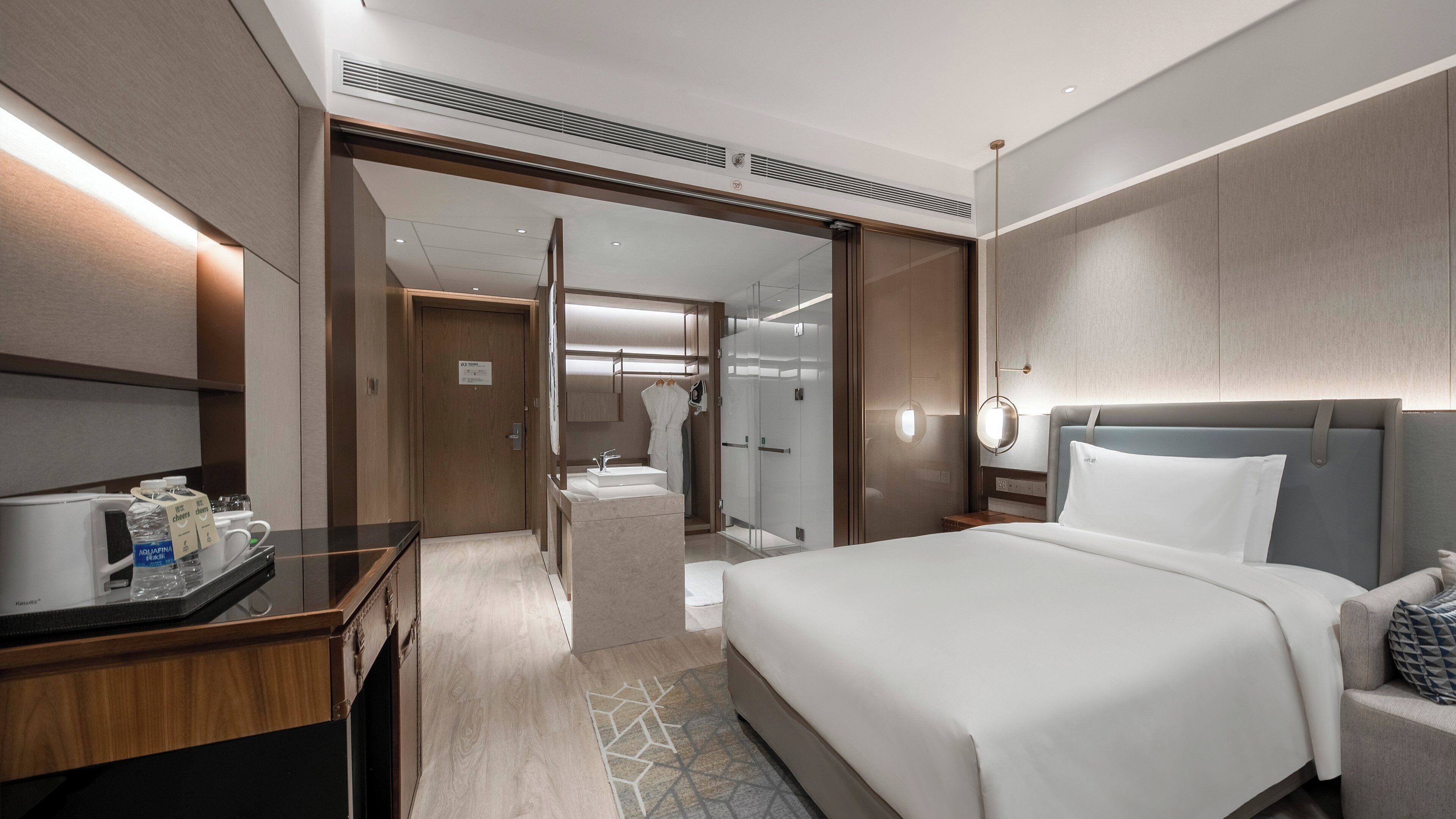 A modern hotel room with a large bed, a desk with a computer, a glass-enclosed shower, and a bathroom with a sink and mirror.
