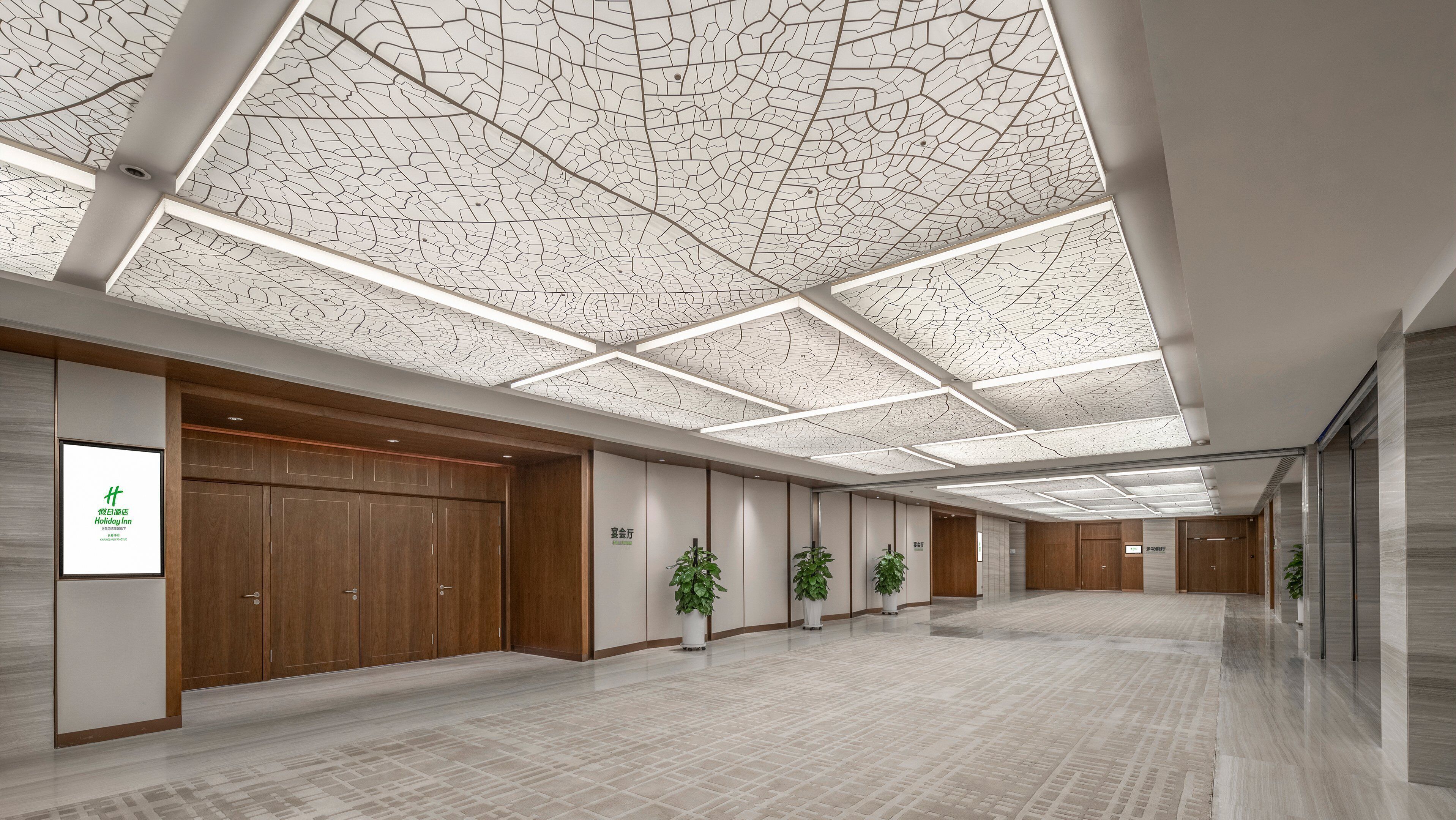 A spacious hotel lobby with a high ceiling featuring a cracked glass design, wooden paneling, and potted plants.