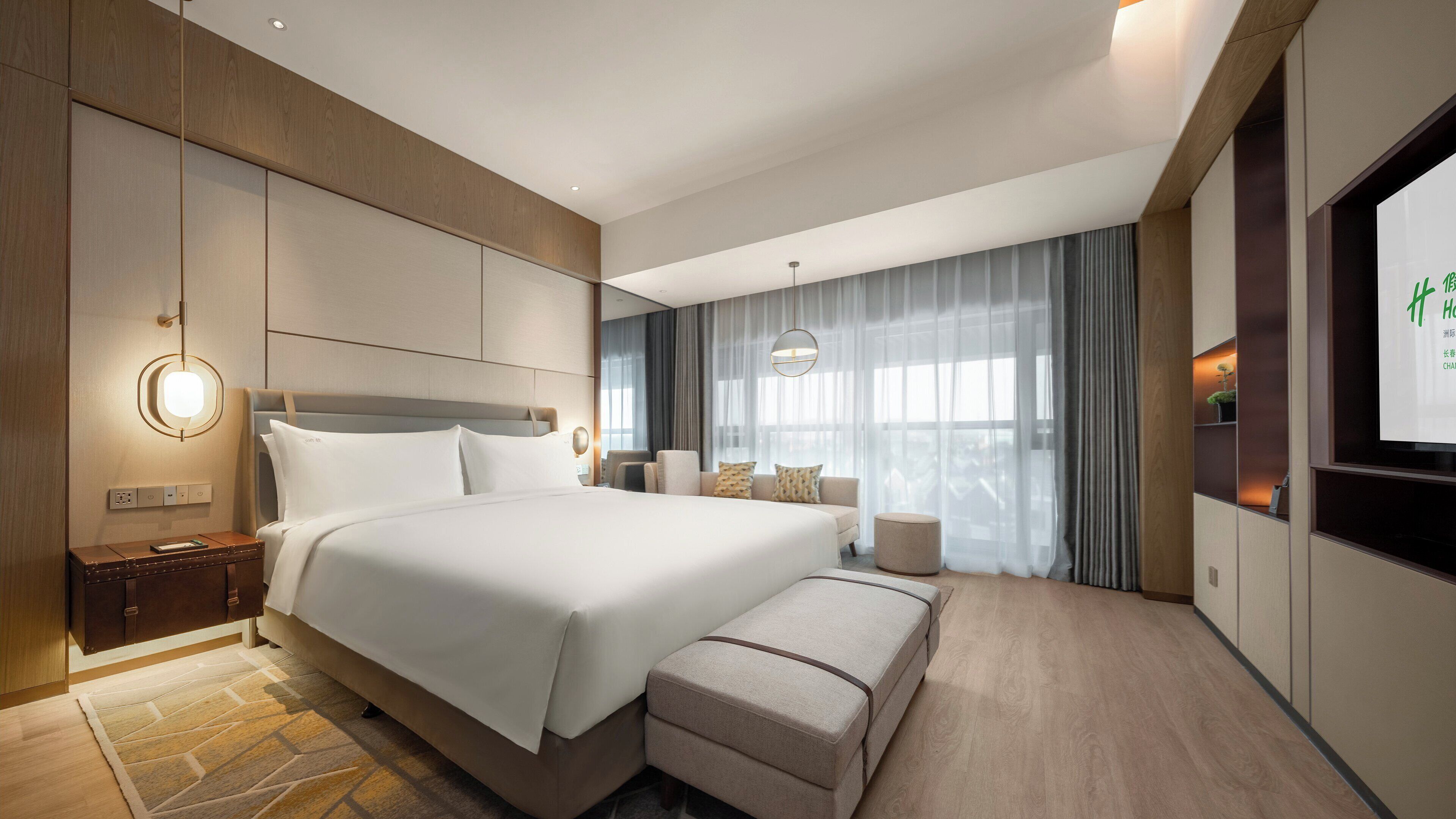 Une chambre d’hôtel moderne avec un grand lit, une table de chevet, un coin salon comprenant un canapé et des fauteuils, une petite table ronde et un téléviseur fixé au mur.