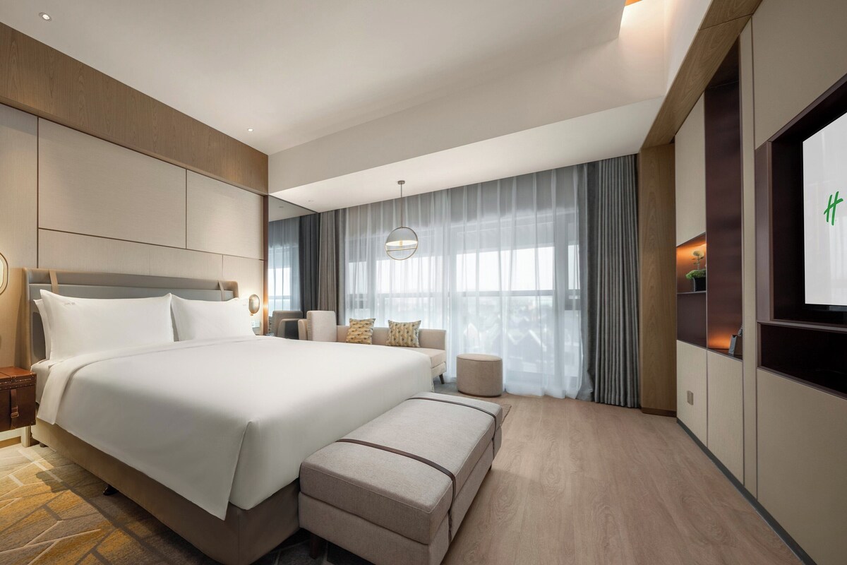 A modern hotel room with a large bed, a bedside table, a seating area with a sofa and armchairs, a small round table, and a TV on the wall.