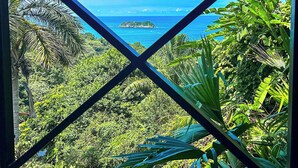 A tropical view through a window with a cross frame, featuring dense greenery and a distant island. - Villa Roca Boutique Resort & Suites-ADULT ONLY (Quepos)