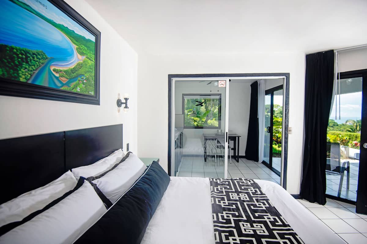Habitación de hotel con cama, un cuadro, un balcón con vistas y una puerta que da a un comedor.