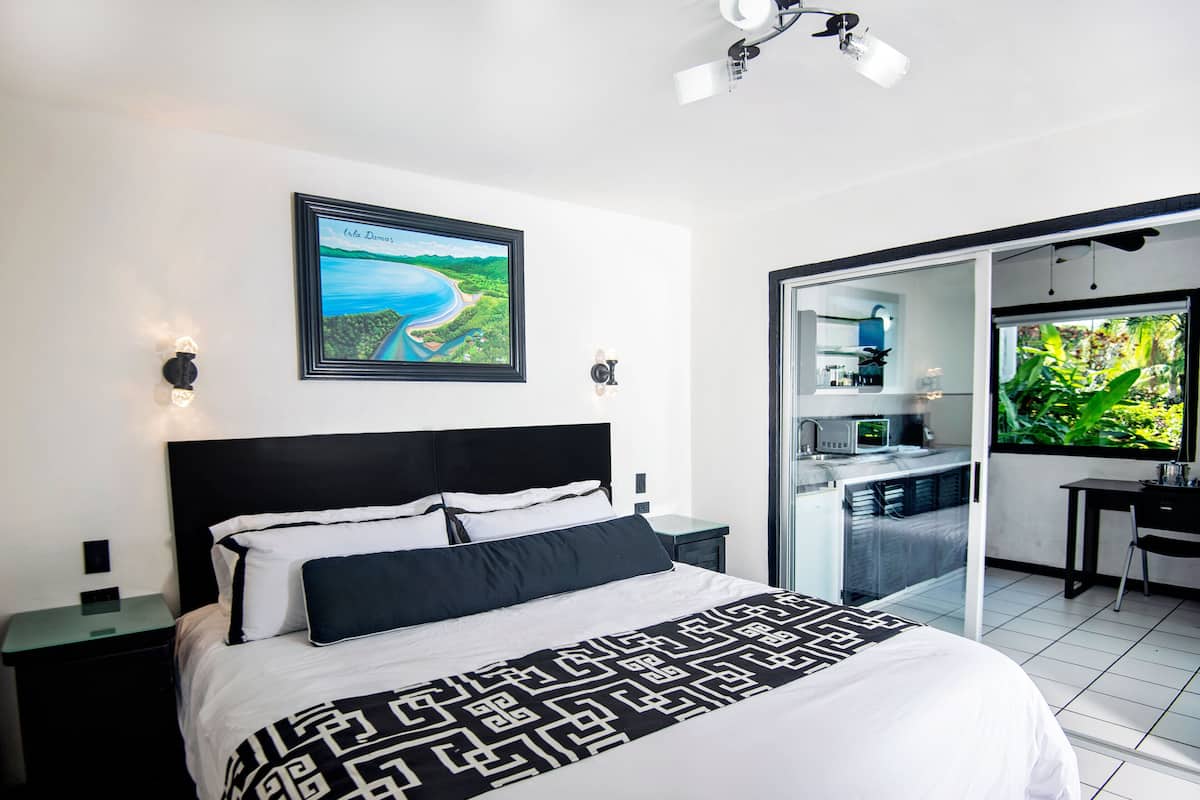 Un dormitorio moderno con una cama grande, vista a una cocina y un cuadro con paisaje en la pared.