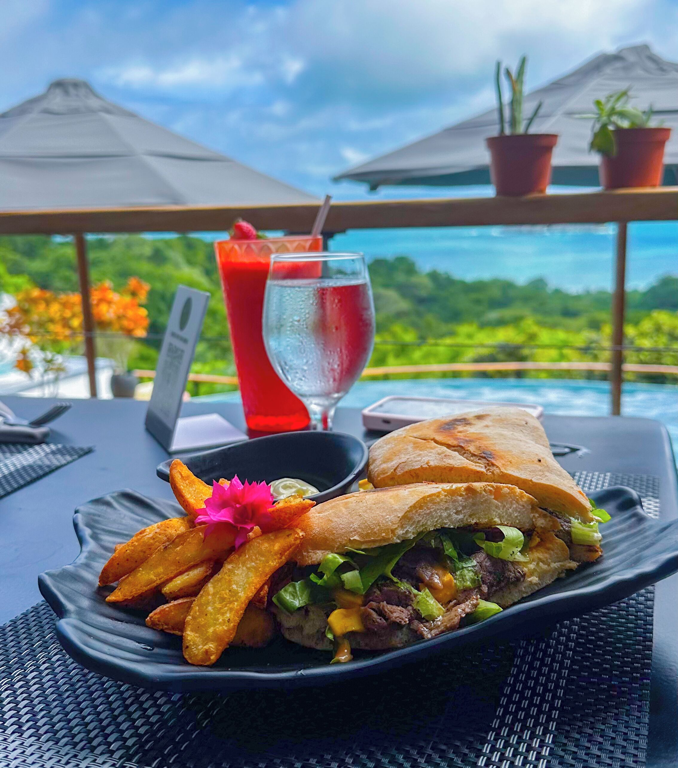 A meal with a sandwich and fries on a plate, a drink, and a laptop on a table with a view of the ocean.