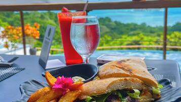 A meal with a sandwich and fries on a plate, a drink, and a laptop on a table with a view of the ocean.